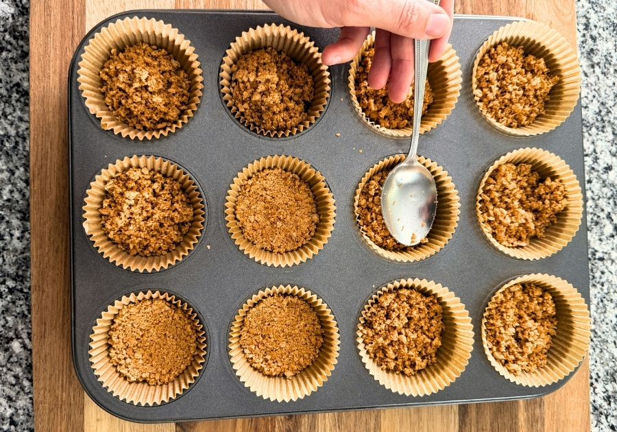 Spoon pressing down on graham cracker mixture to form crust for cheesecakes.